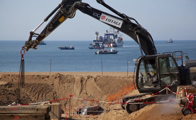 Photo d'un engin de chantier en train de creuser une tranchée sur la plage, et d'un navie câblier en arrière-plan